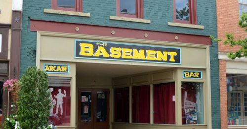 A storefront with a sign reading "The Basement" featuring an arcade and bar, surrounded by greenery and red curtains.