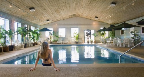 A woman sits by an indoor pool surrounded by plants and lounge chairs, with large windows letting in natural light.