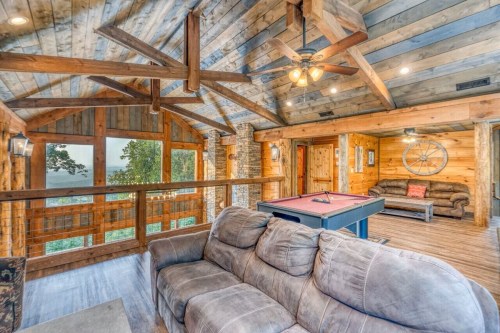 Cozy wooden cabin interior with a pool table, sofa, and large windows showcasing a scenic view.