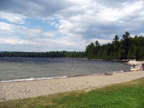 A sandy beach by a lake, surrounded by trees under a cloudy sky. Gentle waves lap at the shore.