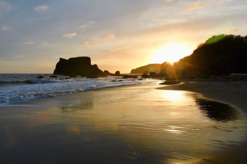 A serene beach at sunset, with gentle waves, rocky formations, and a golden sky reflecting on the wet sand.