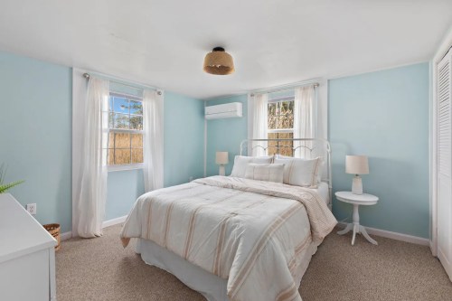 Cozy bedroom with light blue walls, a double bed, and natural light from windows, featuring a nightstand and air conditioning.
