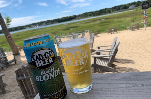 A can and glass of Cape Cod Beach Blonde beer on a wooden table, with a sandy beach and green landscape in the background.