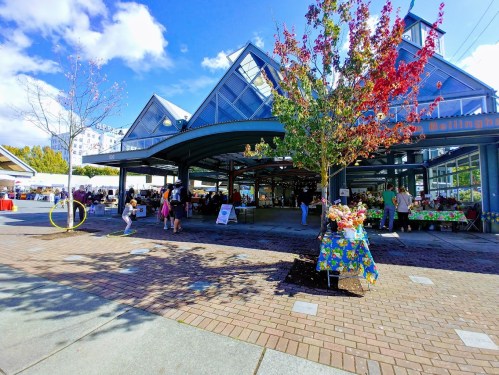 A vibrant market scene with people shopping, colorful stalls, and trees with autumn leaves under a clear blue sky.