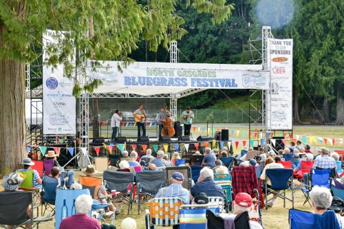 A bluegrass band performs on stage at the North Cascades Bluegrass Festival, with an audience seated in front.