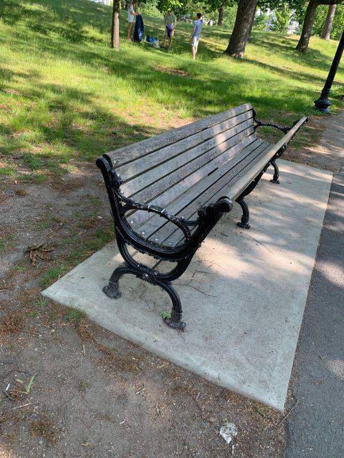 A black metal park bench on a concrete slab, surrounded by grass and trees, with people walking in the background.