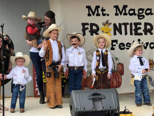 Children dressed as cowboys and cowgirls perform on stage at the Mt. Magan Frontier Festival, with a backdrop and audience.
