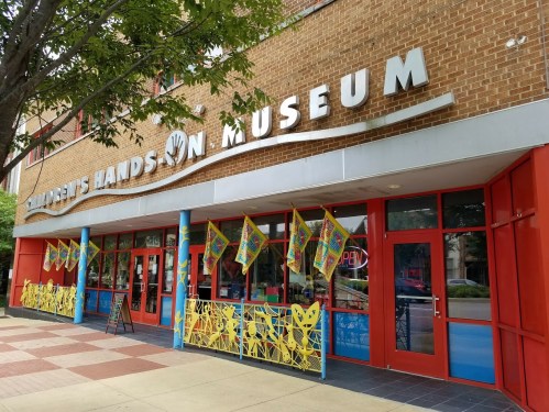 Exterior of the Children's Hands-On Museum, featuring colorful banners and a welcoming entrance with red doors.