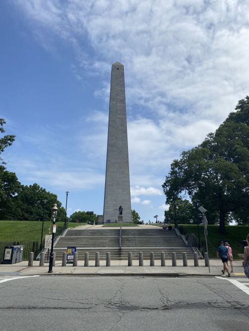 A tall stone monument stands on a grassy hill under a partly cloudy sky, with people walking nearby.