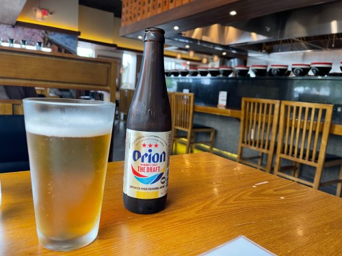 A bottle of Orion beer next to a glass of beer on a wooden table in a restaurant with wooden chairs in the background.