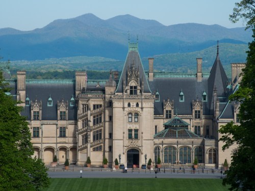 A grand mansion with intricate architecture, set against a backdrop of rolling mountains and clear blue skies.