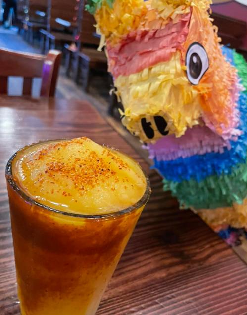 A colorful piñata in the background next to a glass of mango drink topped with chili powder on a wooden table.