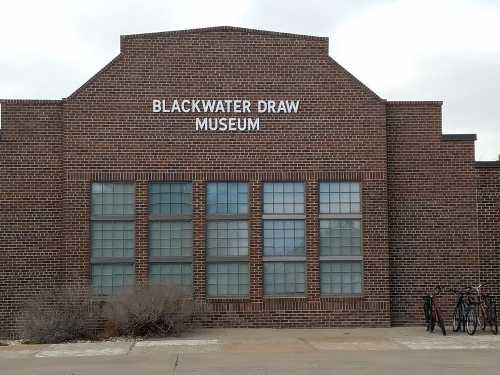 Exterior of the Blackwater Draw Museum, featuring a brick facade and large windows. Bicycles are parked nearby.