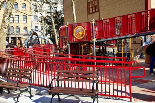 A colorful playground with red structures, slides, and benches in an urban setting.