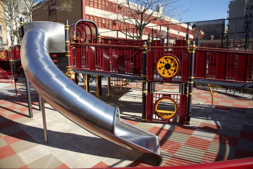 A shiny metal slide on a colorful playground structure with red and yellow accents, surrounded by trees and buildings.