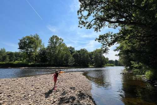 A child walks along a rocky riverbank under a clear blue sky, surrounded by lush green trees.