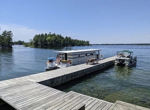 A houseboat docked at a wooden pier on a calm lake, surrounded by lush green trees under a clear blue sky.