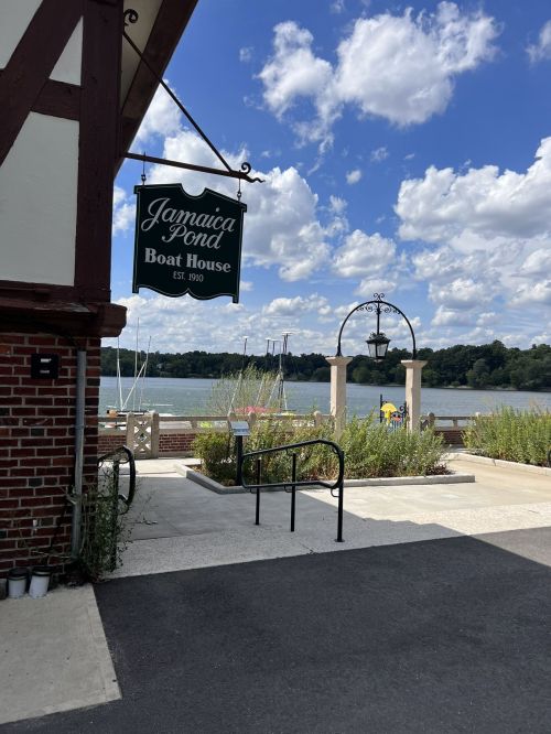 Sign for Jamaica Pond Boat House with a view of the pond and boats under a blue sky with fluffy clouds.