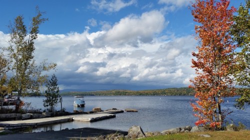 Scenic view of a lake with a dock, colorful trees, and a cloudy sky in the background.