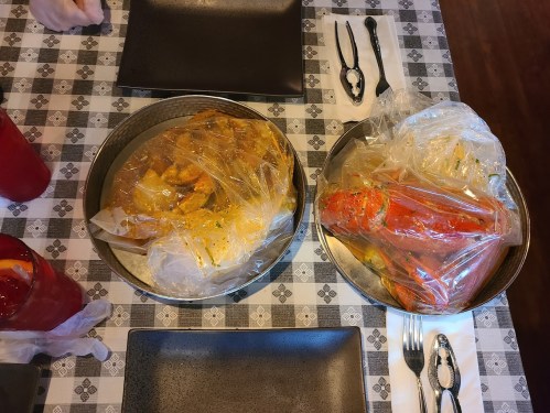 Two metal trays on a checkered tablecloth: one with sealed seafood and the other with a whole lobster in plastic.