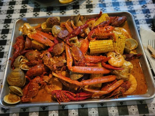 A large tray of seafood featuring crawfish, shrimp, crab, clams, corn, and potatoes, served on a checkered tablecloth.