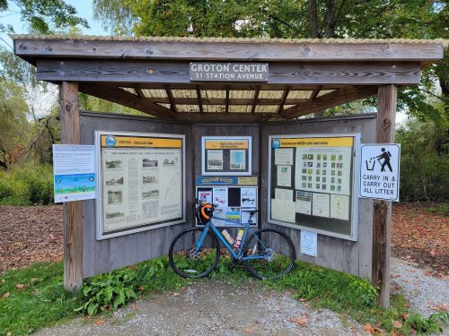 A wooden information kiosk at Groton Center with a bicycle parked in front, featuring various local maps and notices.