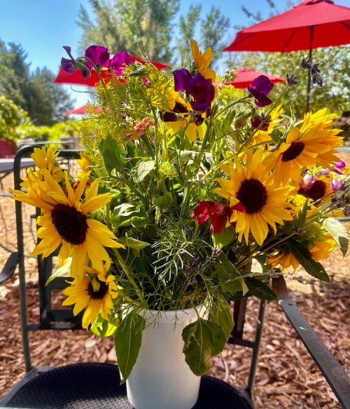 A vibrant bouquet of sunflowers and colorful wildflowers in a white vase, set outdoors with red umbrellas in the background.