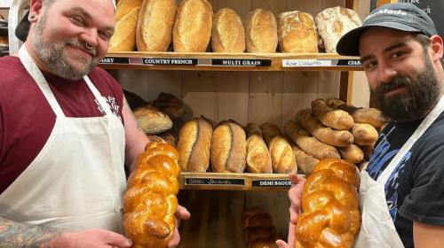 Two bakers proudly hold a braided loaf of bread in front of a shelf filled with various types of bread.