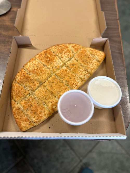 A golden-brown cheesy bread with herbs, cut into squares, served in a pizza box with two dipping sauces.