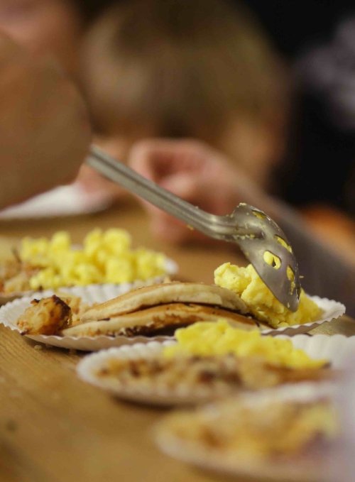 A hand uses a serving utensil to place scrambled eggs on a plate with grilled chicken and tortillas.