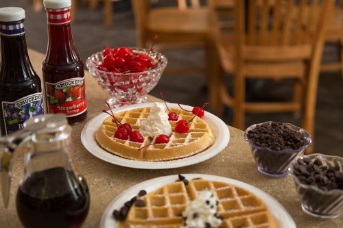 Two plates of waffles topped with whipped cream and cherries, surrounded by syrup and chocolate chips on a table.