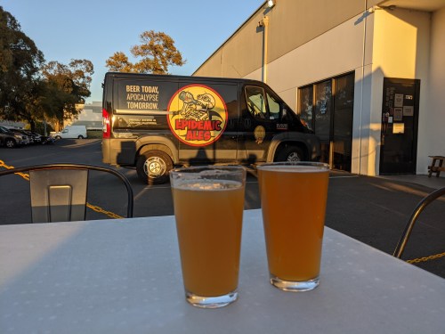 Two glasses of beer sit on a table in front of a van with "Epidemic Ales" branding parked outside a building.