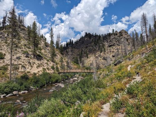 A scenic landscape featuring a wooden bridge over a stream, surrounded by rocky hills and lush greenery under a blue sky.