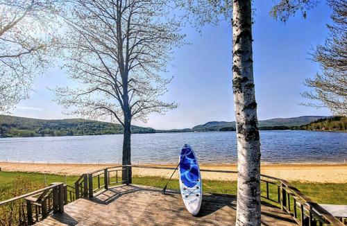 A serene lakeside view with a paddleboard on a wooden deck, surrounded by trees and mountains under a clear blue sky.