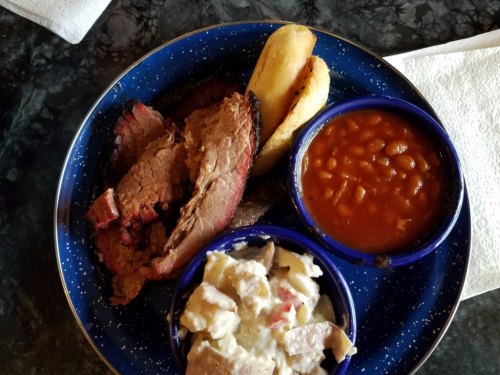 A plate with sliced brisket, baked beans, potato salad, and two baked potatoes on a blue speckled dish.