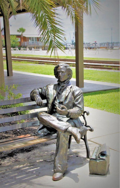 A bronze statue of a seated man in period clothing, resting on a bench with a bag beside him, surrounded by greenery.