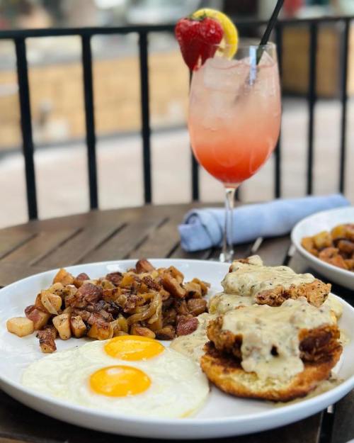 A plate of chicken and biscuits with gravy, two sunny-side-up eggs, and seasoned potatoes, alongside a fruity cocktail.