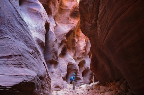 A hiker explores a narrow, colorful canyon with smooth, layered rock formations and sunlight filtering through.