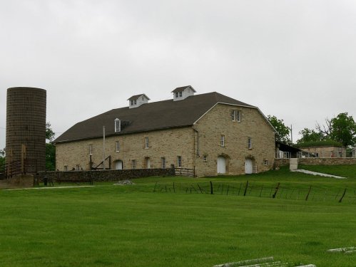 A large stone barn with a silo, set in a green field under a cloudy sky.