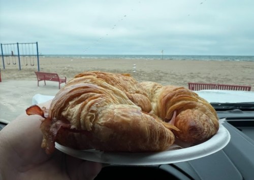 A hand holding a plate with croissants in front of a beach view on a cloudy day.