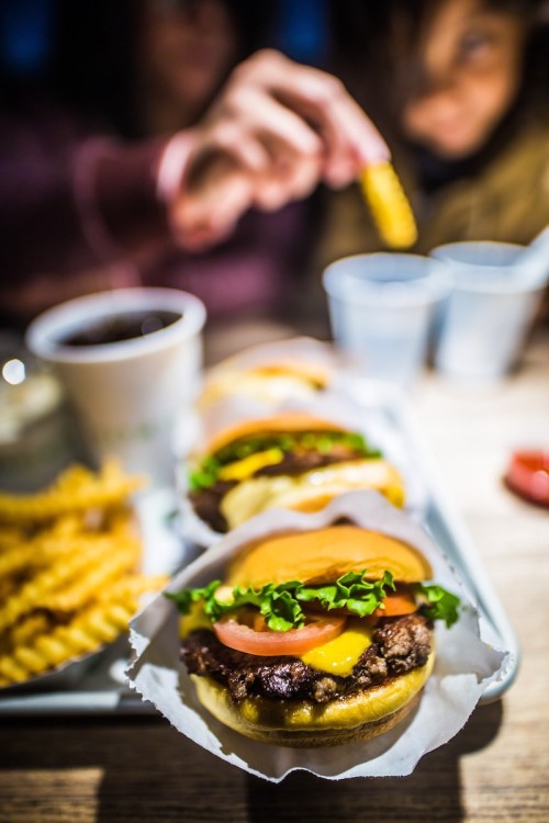 A close-up of burgers with lettuce, tomato, and cheese, accompanied by crinkle fries and drinks in the background.