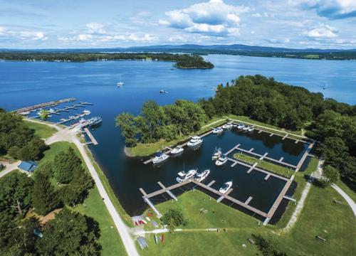 Aerial view of a marina with boats docked, surrounded by green trees and a calm lake under a blue sky with clouds.