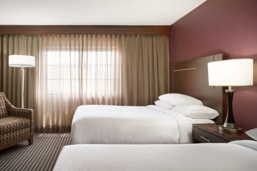 A hotel room featuring two beds, a patterned chair, and large windows with sheer curtains.