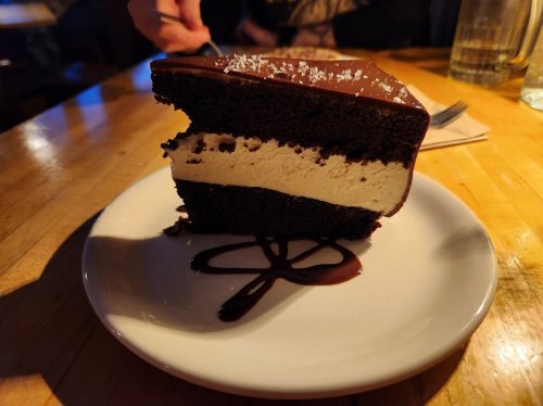 A slice of chocolate cake with a creamy filling, drizzled with chocolate sauce, on a white plate.