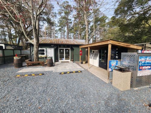 A small green building with a wooden deck, surrounded by trees and gravel, featuring outdoor seating and signage.