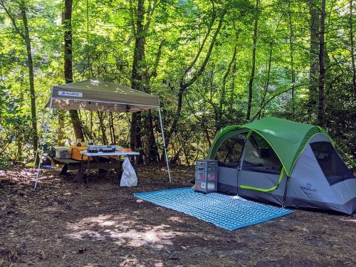A campsite with a green tent, a canopy over a picnic table, and a blue mat on the forest floor surrounded by trees.