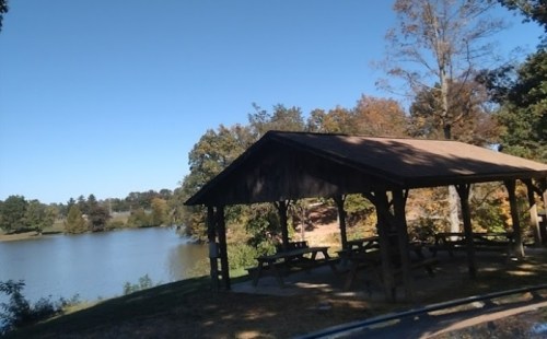 A wooden pavilion by a calm lake surrounded by trees with autumn foliage under a clear blue sky.