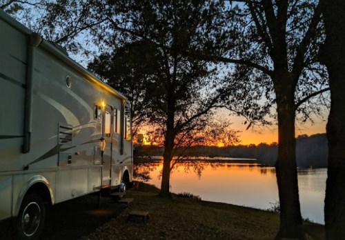 A camper parked by a tranquil lake at sunset, with trees framing the serene scene.