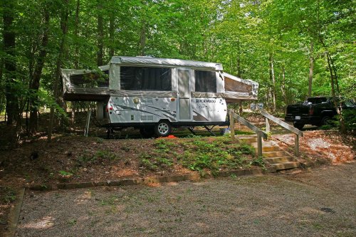 A pop-up camper parked on a wooded lot, surrounded by trees and a gravel driveway.