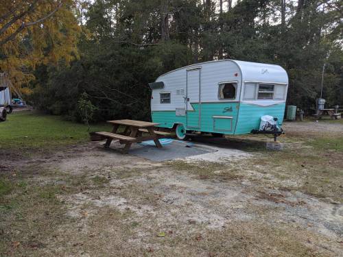 A vintage teal and white camper parked near a picnic table in a grassy area surrounded by trees.
